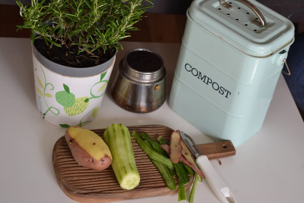 A flower pot with rosemary and some vegetables next to a composting box