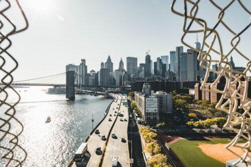 View of the Manhattan skyline and Brooklyn Bridge with traffic along the waterfront highway, captured through a metal fence.