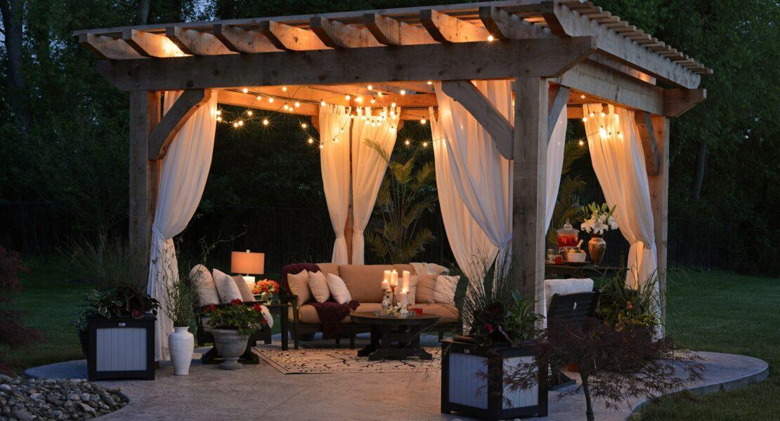 Wooden backyard pergola with white curtains, cozy seating, string lights, and landscaping creating a peaceful outdoor oasis.