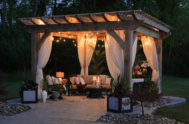 Wooden backyard pergola with white curtains, cozy seating, string lights, and landscaping creating a peaceful outdoor oasis.