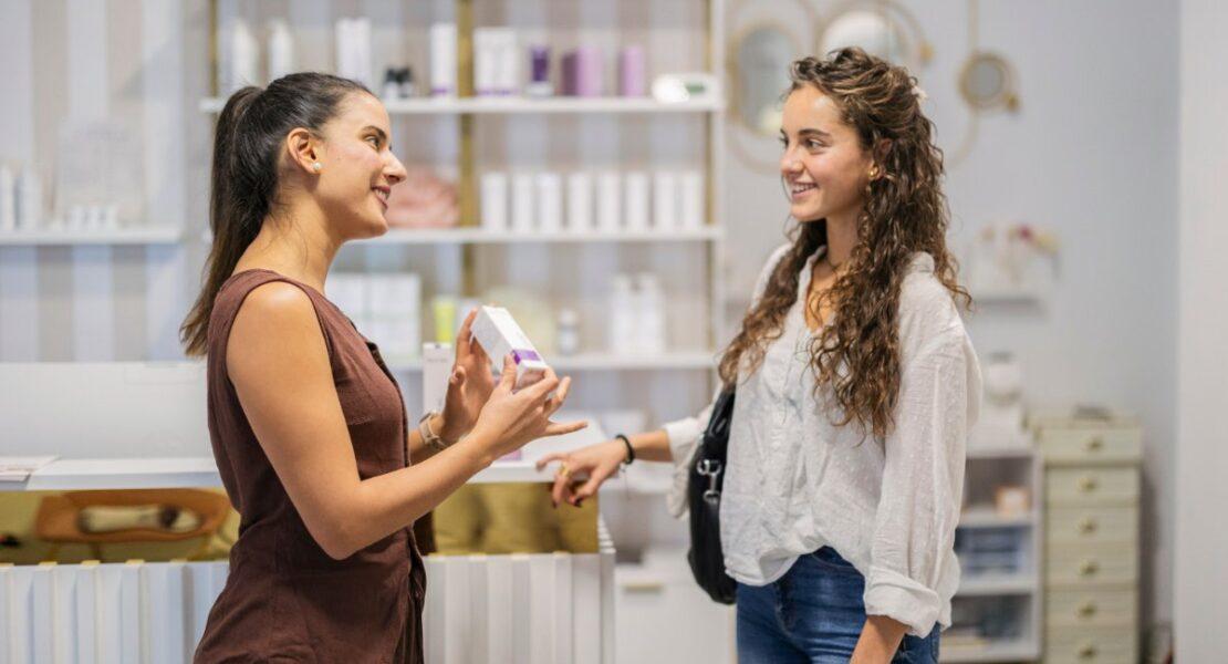 Beauty consultant showing lash and brow aftercare products to a customer at a modern retail display.