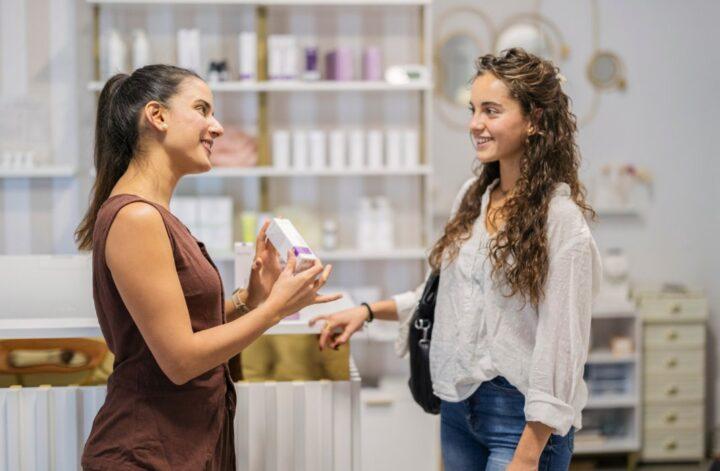 Beauty consultant showing lash and brow aftercare products to a customer at a modern retail display.