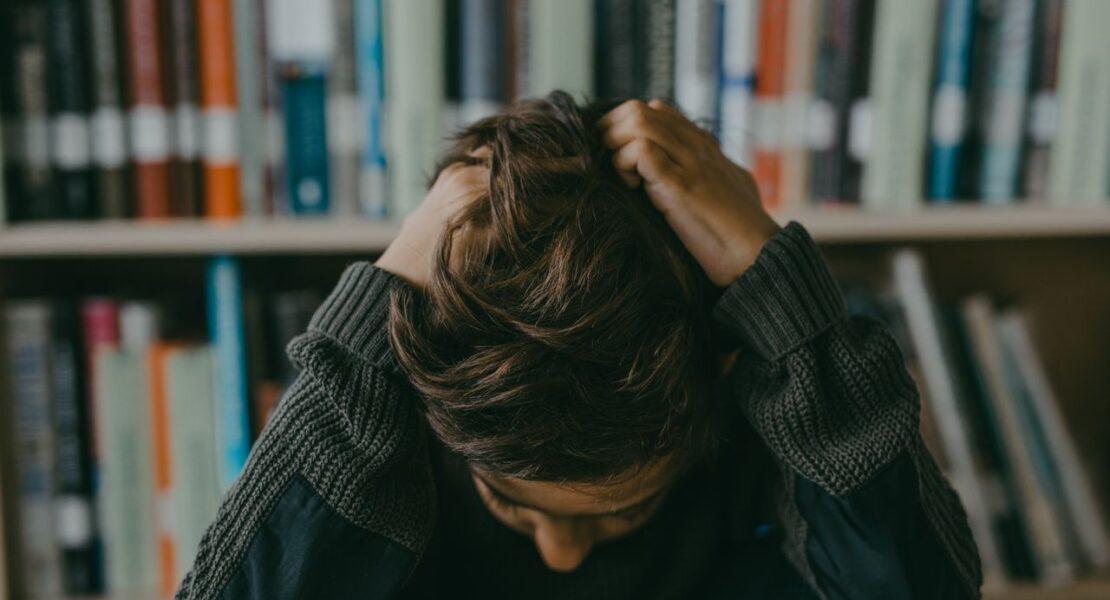 Teen sitting with head down and hands in hair in a library, illustrating signs of anxiety that parents may overlook.