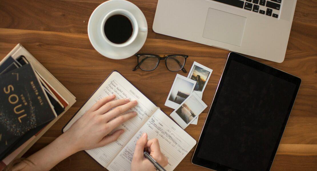 Person journaling beside a laptop, tablet, photos, and coffee while crafting a digital story.