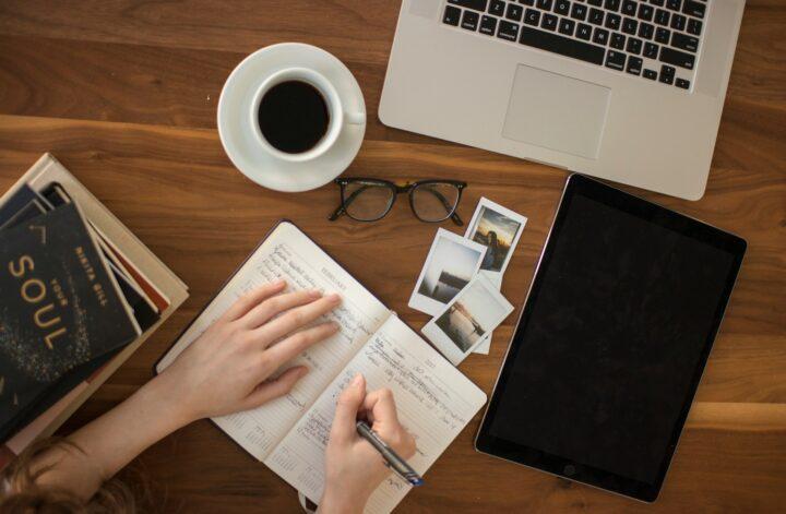 Person journaling beside a laptop, tablet, photos, and coffee while crafting a digital story.