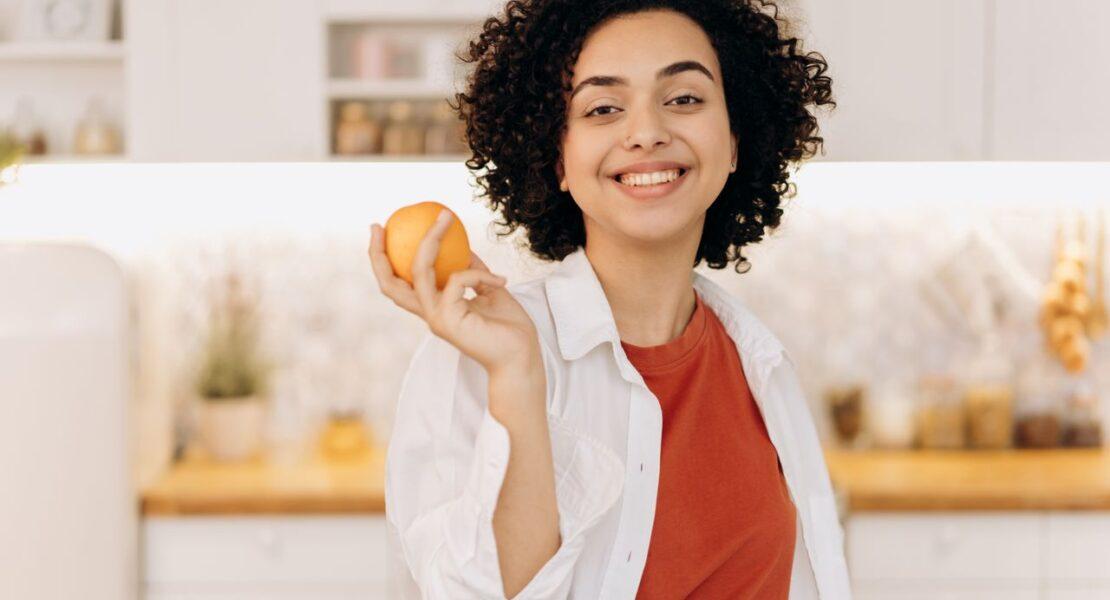 Smiling woman holding an orange in a kitchen while practicing a healthy daily habit