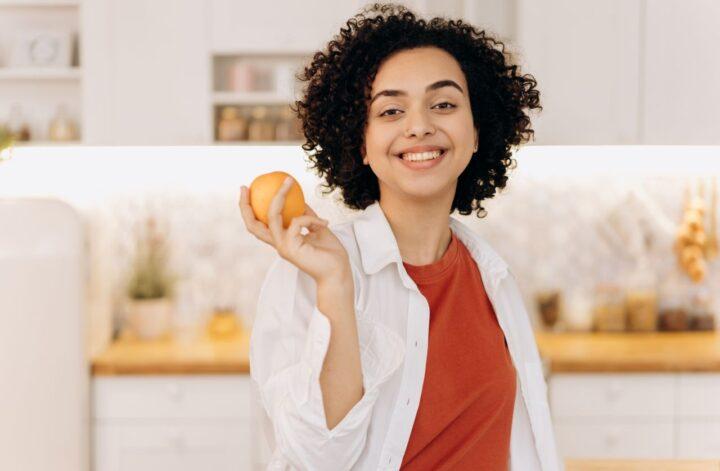 Smiling woman holding an orange in a kitchen while practicing a healthy daily habit