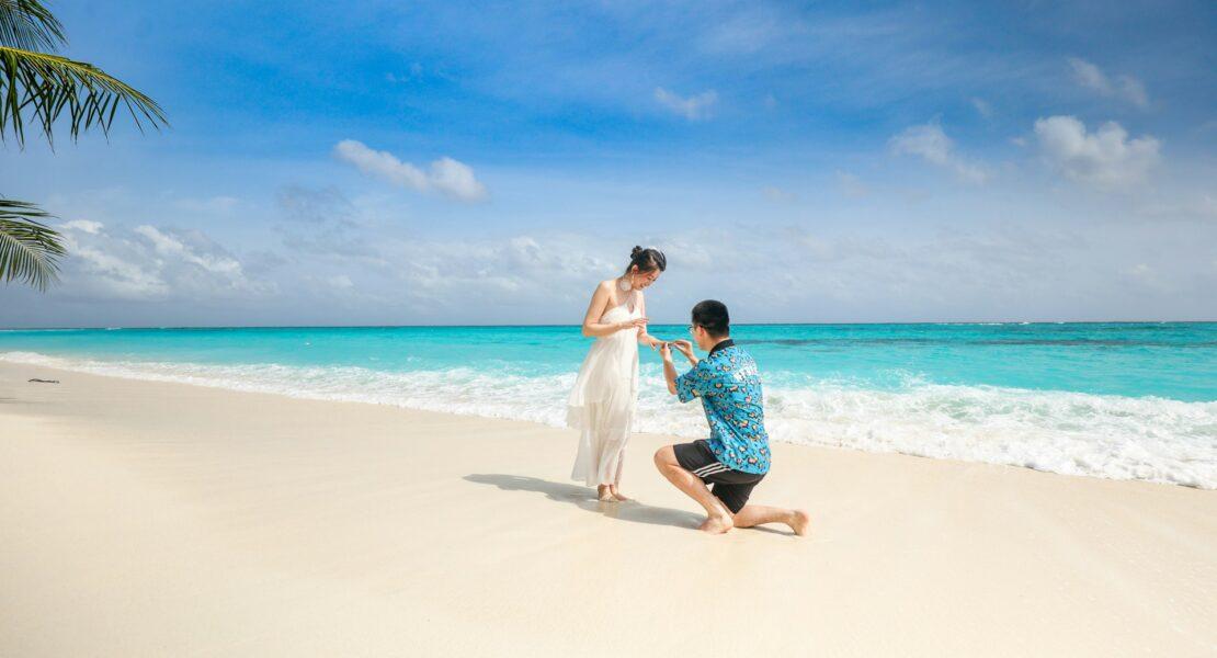 Couple celebrating a beach proposal as the man kneels with a ring beside the ocean.