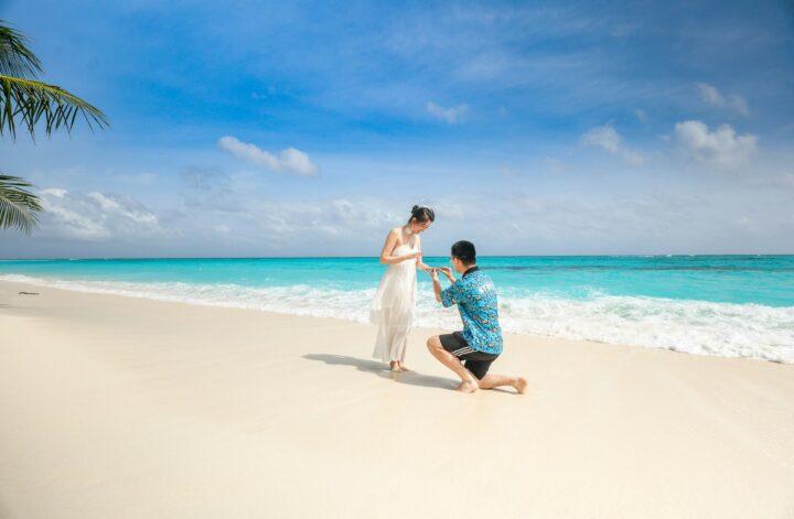 Couple celebrating a beach proposal as the man kneels with a ring beside the ocean.