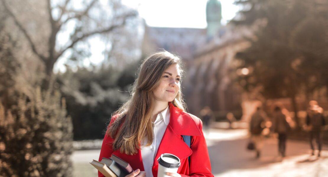 Woman in a red coat carrying books and coffee while enjoying a sunny outdoor setting.