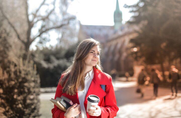 Woman in a red coat carrying books and coffee while enjoying a sunny outdoor setting.