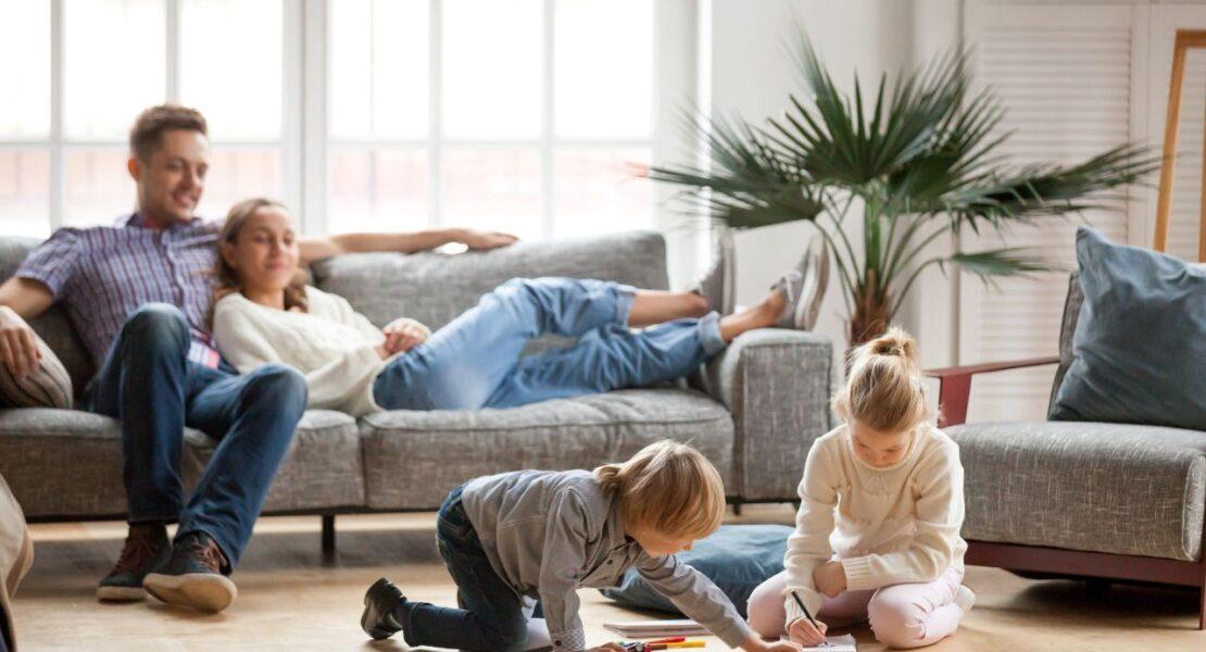 Parents relax on a sofa while two children draw on the living room floor.