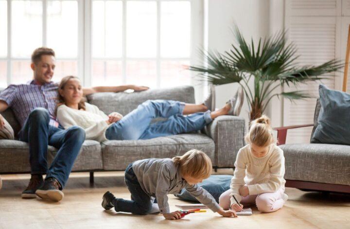 Parents relax on a sofa while two children draw on the living room floor.