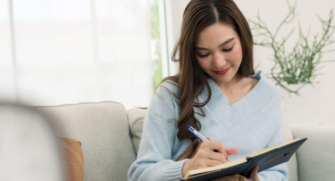 Young woman writing in a notebook while relaxing on a sofa, an example of a screen-free activity for young adults.