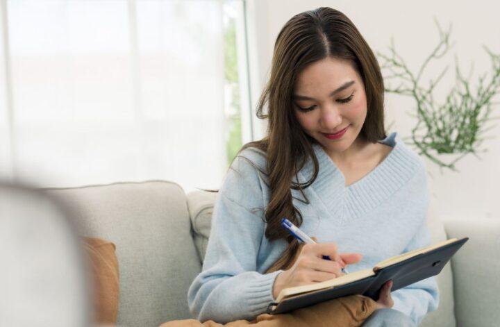 Young woman writing in a notebook while relaxing on a sofa, an example of a screen-free activity for young adults.